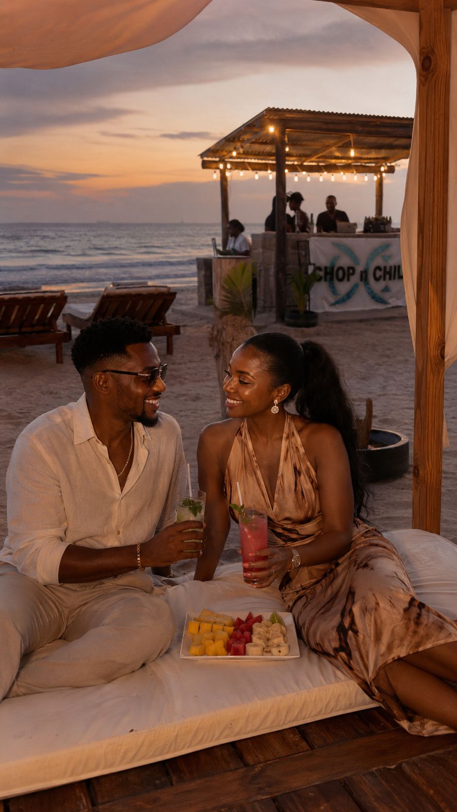 A relaxed couple enjoying drinks and fruit on a beach cabana at sunset